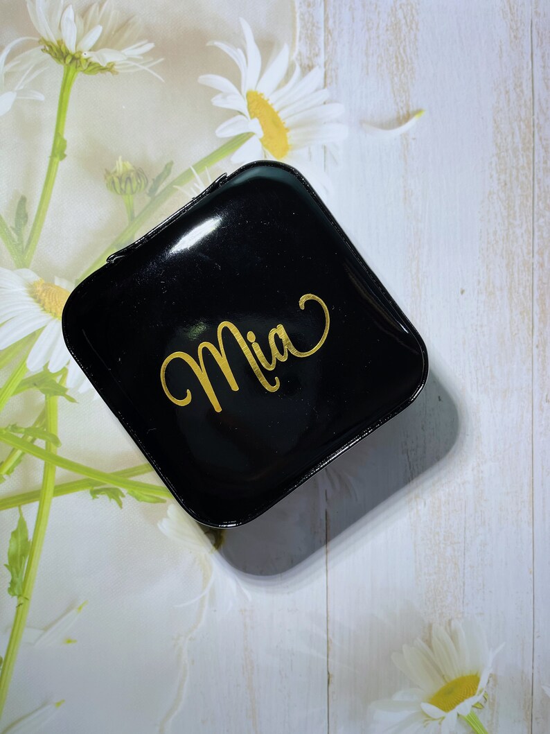 Pu&ograve; includere: Un portagioie nero, quadrato e lucido con il nome "Mia" in oro. La scatola &egrave; chiusa e poggia su uno sfondo floreale bianco e verde chiaro con fiori di margherita.