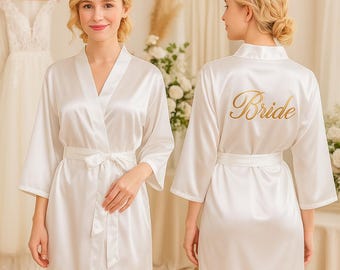 May include: A white satin bridal robe with a matching belt. The robe has a V-neck and three-quarter sleeves. The word "Bride" is printed in gold script on the back. The robe is knee-length.
