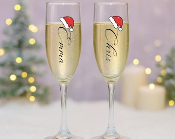 May include: Two champagne flutes filled with a bubbly, golden liquid. Each glass is decorated with a small Santa hat and a name in elegant script: "Emma" and "Chris". The background features soft bokeh lights and a Christmas tree.