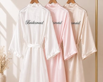 May include: Three satin bridesmaid robes in white, pink, and champagne hang on a gold rack. The white robe has the word "Bridesmaid" printed on the back in black script. Each robe has a matching belt.