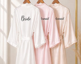 May include: Three satin robes hanging on a gold rack. The robes are white, pink, and champagne-colored. The white robe has the word "Bride" printed on the back, while the pink and champagne robes say "'lesmaid".