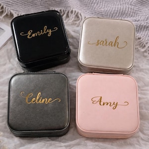 Pu&ograve; includere: Quattro portagioie quadrati in nero, beige, grigio e rosa. Ogni custodia ha un nome in oro: Emily, Sarah, Celine e Amy. Le custodie sono su una superficie bianca e grigia strutturata.