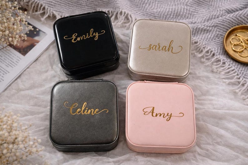 Pu&ograve; includere: Quattro portagioie quadrati in nero, beige, grigio e rosa. Ogni custodia ha un nome in oro: Emily, Sarah, Celine e Amy. Le custodie sono su una superficie bianca e grigia strutturata.