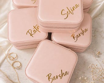 May include: A collection of six pink jewelry cases with gold lettering, each bearing a different name. The cases are square with a zipper closure and are arranged on a white, textured surface. Gold necklaces and rings are visible.