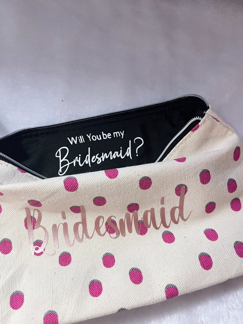 May include: A white canvas zippered pouch with pink polka dots. The pouch is open and the inside lining is black. The text "Will You be my Bridesmaid?" is printed in white on the black lining. The text "Bridesmaid" is printed in pink on the front of the pouch.