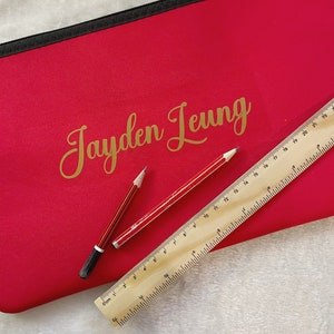 May include: A bright pink pencil case with a black zipper and the name "Jayden Leung" written in gold lettering. Two pencils and a wooden ruler are lying on top of the case.