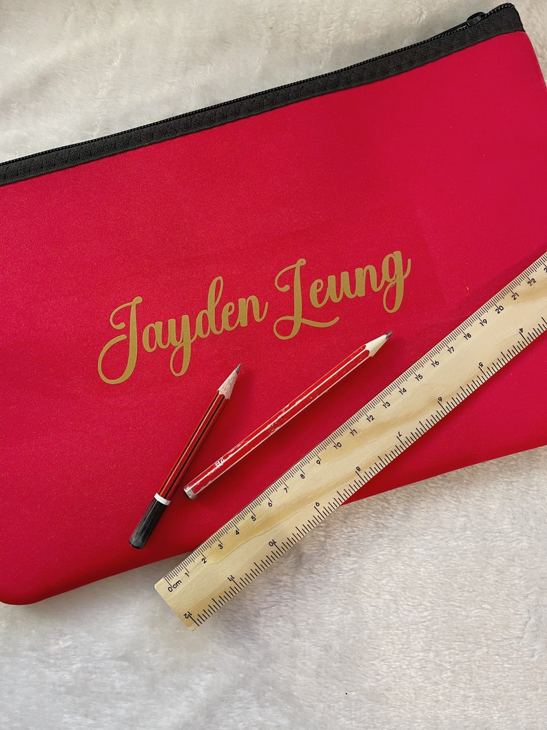 May include: A bright pink pencil case with a black zipper and the name "Jayden Leung" written in gold lettering. Two pencils and a wooden ruler are lying on top of the case.