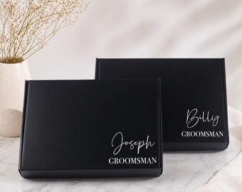 May include: Two black gift boxes with white text. One box reads "Joseph GROOMSMAN" and the other reads "Billy GROOMSMAN". The boxes are on a marble surface, with a vase of dried flowers in the background.