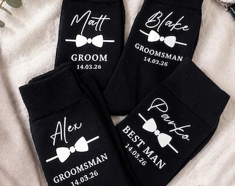 May include: Four black socks with white text and bow tie graphics. The socks are personalized with names, titles, and dates: Matt, Groom, 14.03.26; Blake, Groomsman, 14.03.26; Alex, Groomsman, 14.03.26; and Parko, Best Man, 14.03.26.
