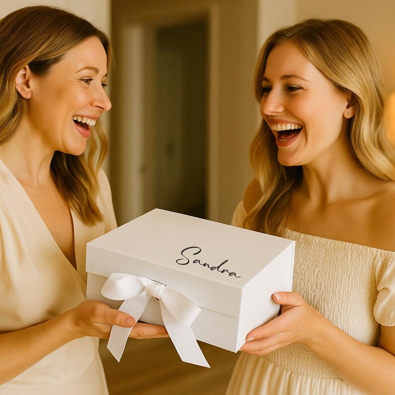 May include: A white gift box with a satin ribbon bow, held by two people. The box has the name "Sandra" written on the top in a cursive font. The people are smiling and appear to be excited.