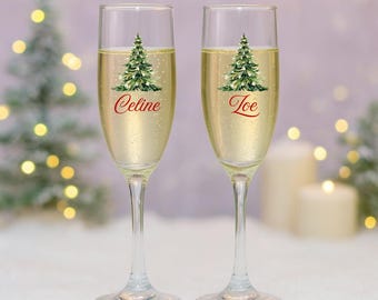 May include: Two champagne flutes filled with a bubbly, golden liquid. Each glass features a Christmas tree design and a name in red script: "Celine" and "Joe". The background includes soft bokeh lights and a snowy surface, suggesting a festive holiday setting.