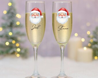 May include: Two champagne flutes filled with a sparkling beverage. Each glass features a Santa Claus face and a name in cursive script. The background includes a blurred Christmas tree and soft, glowing lights, creating a festive holiday scene.