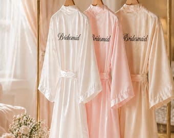 May include: Three satin bridesmaid robes in white, pink, and champagne hang on a gold rack. Each robe has the word "Bridesmaid" printed on the back in black script. A pair of white high heels and a bouquet of flowers are in the foreground.
