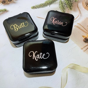 Pu&ograve; includere: Tre portagioie neri quadrati con nomi personalizzati in corsivo oro, rosa e bianco: "Brett", "Karen" e "Kate". Le scatole sono lucide e con cerniera, suggerendo un tema di viaggio o conservazione.