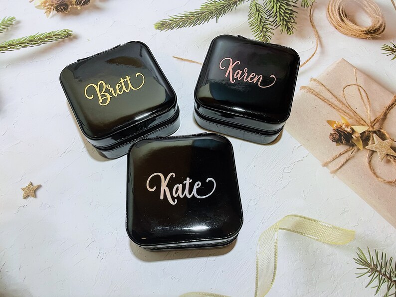 Pu&ograve; includere: Tre portagioie neri quadrati con nomi personalizzati in corsivo oro, rosa e bianco: "Brett", "Karen" e "Kate". Le scatole sono lucide e con cerniera, suggerendo un tema di viaggio o conservazione.