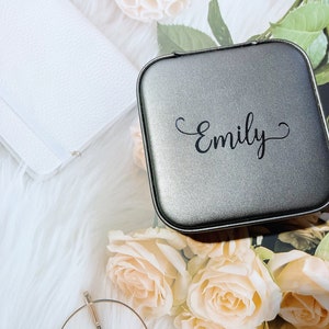 Pu&ograve; includere: Un portagioie quadrato grigio con il nome "Emily" in corsivo nero. La scatola &egrave; circondata da rose color pesca, un portafoglio bianco e una superficie bianca e soffice.