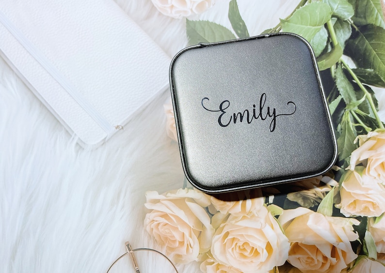 Pu&ograve; includere: Un portagioie quadrato grigio con il nome "Emily" in corsivo nero. La scatola &egrave; circondata da rose color pesca, un portafoglio bianco e una superficie bianca e soffice.
