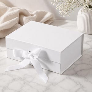 May include: A white gift box with a white ribbon tied in a bow. The box is rectangular and has a magnetic closure. The background includes a white vase with flowers and a white cloth.