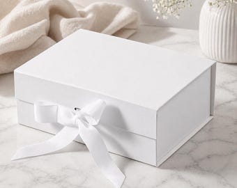 May include: A white gift box with a matching white ribbon tied in a bow. The box is rectangular and has a magnetic closure. The background includes a white vase with flowers and a white cloth.