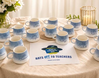 May include: A collection of blue and white gradient mugs with names written on them, arranged on a beige tablecloth. A sign that reads "Hats Off To Teachers" is in the center. The mugs are for World Teachers' Day.
