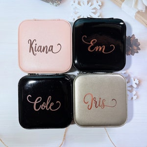 Pu&ograve; includere: Quattro custodie quadrate e personalizzate per lenti a contatto in rosa, nero e argento. Ogni custodia ha un nome in corsivo: Kiana, Em, Cole e Iris. Le custodie sono disposte su una superficie bianca con decorazioni a fiocchi di neve e pigne.