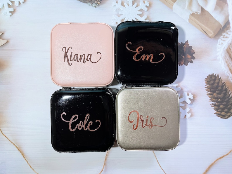 Pu&ograve; includere: Quattro custodie quadrate e personalizzate per lenti a contatto in rosa, nero e argento. Ogni custodia ha un nome in corsivo: Kiana, Em, Cole e Iris. Le custodie sono disposte su una superficie bianca con decorazioni a fiocchi di neve e pigne.