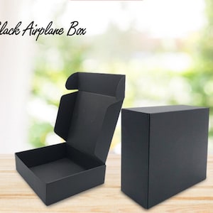 May include: Two black cardboard boxes, one open and one closed. The boxes are on a wooden surface. The text "Black Airplane Box" is visible at the top of the image.