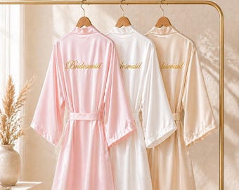 May include: Three satin bridesmaid robes in pink, white, and beige hang on a gold rack. Each robe has a matching belt and the word "Bridesmaid" or "desmaid" embroidered in gold script on the back. The robes are displayed on wooden hangers.