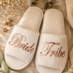 May include: Two white, open-toe slippers with rose gold script reading "Bride" and "Tribe". The slippers are made of a soft, plush material. Ideal for a bridal party or as a wedding gift.