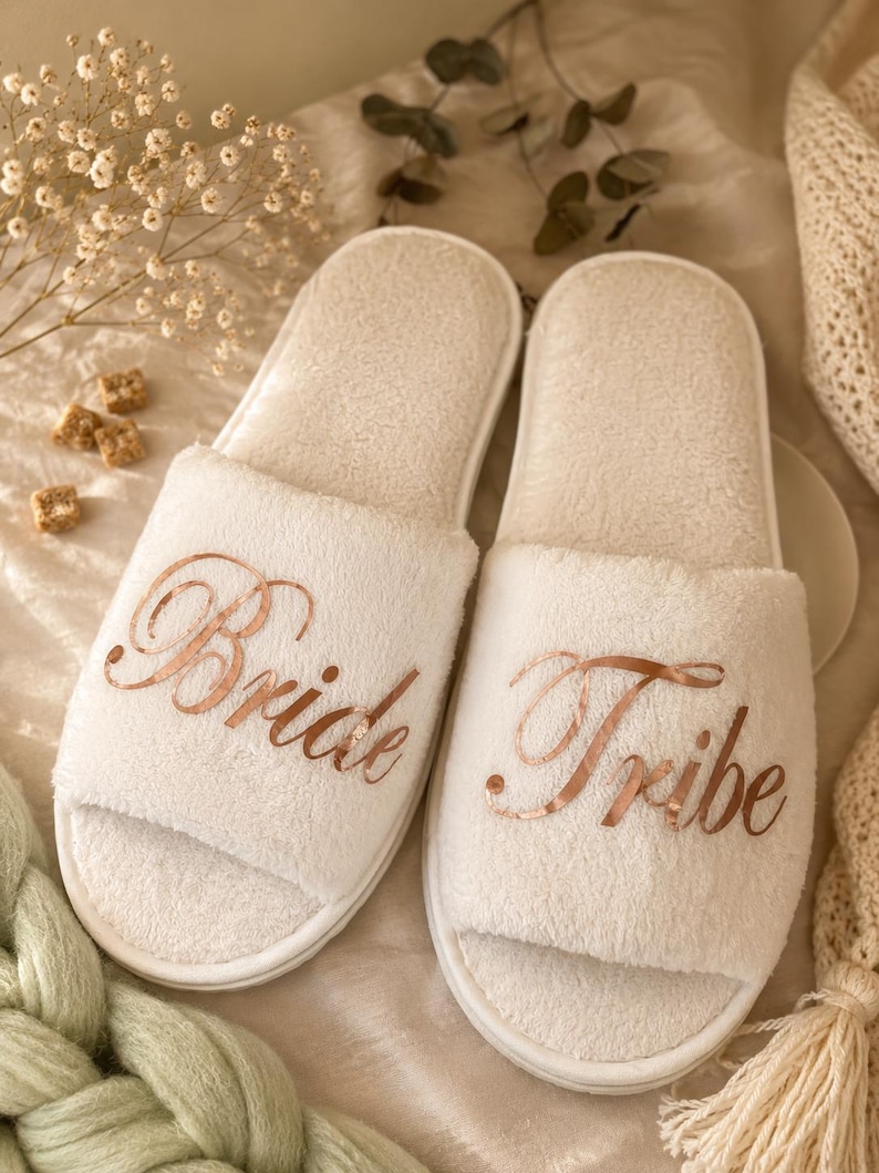 May include: Two white, open-toe slippers with rose gold script reading "Bride" and "Tribe". The slippers are made of a soft, plush material. Ideal for a bridal party or as a wedding gift.