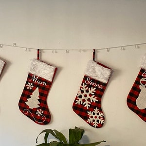May include: Four red and black plaid Christmas stockings with white fur trim, personalized with the names "Steve", "Mum", "Sienna", and "Bayden". Each stocking features a different white embroidered design: a reindeer, a Christmas tree, a snowflake, and a reindeer.