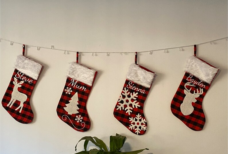 May include: Four red and black plaid Christmas stockings with white fur trim, personalized with the names "Steve", "Mum", "Sienna", and "Bayden". Each stocking features a different white embroidered design: a reindeer, a Christmas tree, a snowflake, and a reindeer.