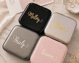 May include: Four square jewelry boxes in black, silver, gray, and pink, each with a name in elegant gold or white script. A card with flowers and a small dish with jewelry are also visible.