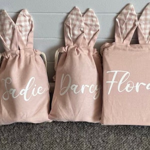 May include: Five pink and green bunny-shaped drawstring bags with white lettering. The bags are personalized with the names Indie, Sadie, Darcy, Flora, and Leo.