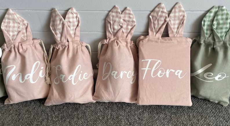 May include: Five pink and green bunny-shaped drawstring bags with white lettering. The bags are personalized with the names Indie, Sadie, Darcy, Flora, and Leo.