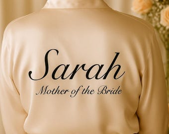 May include: Cream-colored satin robe with the name "Sarah" and the text "Mother of the Bride" in elegant black script. The robe has a wrap-around design with a matching belt, perfect for bridal parties.