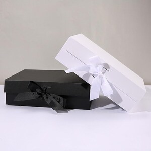 May include: Two rectangular gift boxes, one black and one white, are displayed. The black box is closed with a black ribbon tied in a bow. The white box is open, with a white ribbon tied in a bow. The boxes are on a white surface.