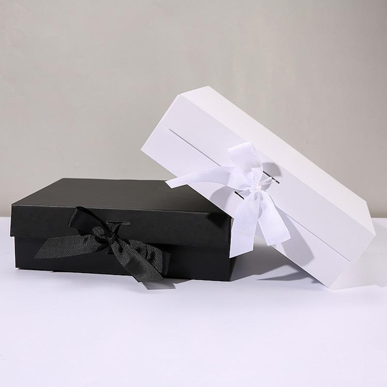 May include: Two rectangular gift boxes, one black and one white, are displayed. The black box is closed with a black ribbon tied in a bow. The white box is open, with a white ribbon tied in a bow. The boxes are on a white surface.