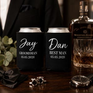 May include: Two black foam can coolers featuring white printed text. The left cooler displays the name Jay, the title GROOMSMAN, and the date 05.05.2028. The right cooler displays the name Dan, the title BEST MAN, and the date 05.05.2028.