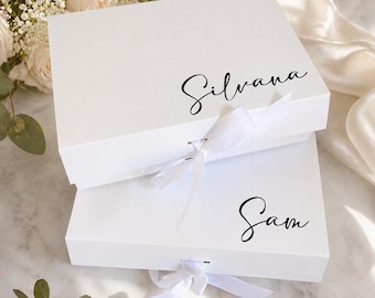 May include: Two white gift boxes, one atop the other, with the names "Silvana" and "Sam" in elegant black script. Each box is tied with a white ribbon. The boxes are surrounded by flowers and greenery, suggesting a celebratory occasion.
