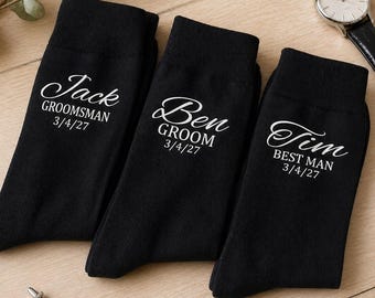 May include: Three black socks with white text, personalized for a wedding. The socks are labeled "Jack GROOMSMAN 3/4/27", "Ben GROOM 3/4/27", and "Tim BEST MAN 3/4/27". Also visible are a watch and cufflinks.