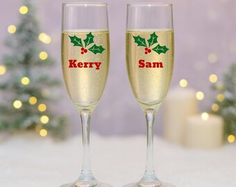 May include: Two champagne flutes filled with a bubbly beverage. Each glass is decorated with a green holly and red berry design and a name in red text. The background features a blurred Christmas tree and lights.