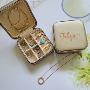 Pu&ograve; includere: Un portagioie beige con il nome "Taliya" in corsivo color oro rosa. La scatola aperta mostra anelli, bracciali e collane. &Egrave; visibile una collana d'oro con un pendente circolare. Il portagioie &egrave; su una superficie bianca.