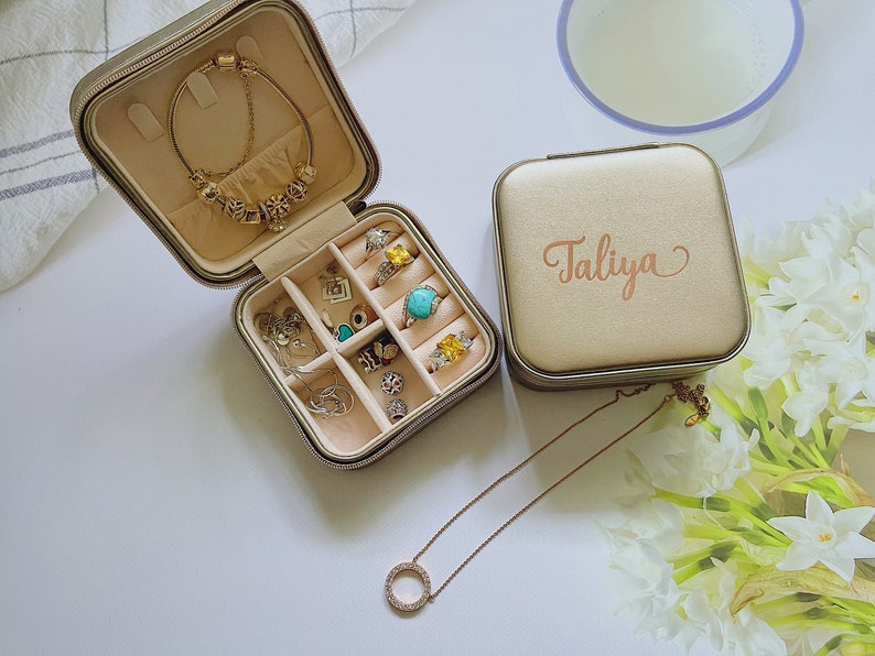 Pu&ograve; includere: Un portagioie beige con il nome "Taliya" in corsivo color oro rosa. La scatola aperta mostra anelli, bracciali e collane. &Egrave; visibile una collana d'oro con un pendente circolare. Il portagioie &egrave; su una superficie bianca.