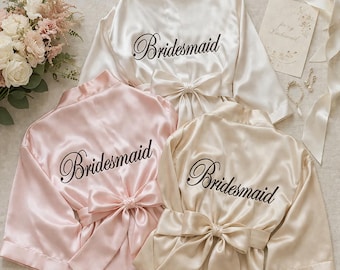 May include: Three satin bridesmaid robes in ivory, blush pink, and champagne. Each robe has a matching sash and the word "Bridesmaid" embroidered on the back in elegant script. A bouquet and accessories are in the background.