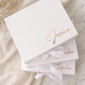Può includere: Tre scatole regalo bianche impilate, ognuna con un nome in elegante script oro rosa: Jessica, Kaylee e Sophie. Ogni scatola è legata con un nastro di raso bianco. Le scatole sono su una superficie di tessuto dai toni neutri.