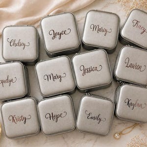 Pu&ograve; includere: Scatole portagioie quadrate argentate con nomi come "Chelsey" e "Mary" scritti in corsivo. Le scatole sono disposte su una superficie chiara, suggerendo un regalo personalizzato o una soluzione di archiviazione.