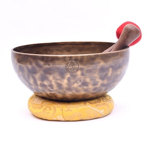 May include: A bronze-colored metal singing bowl with a mottled, dark patina finish and a small circular symbol engraved on the side. The bowl rests on a round, yellow fabric cushion with a gold pattern and includes a wooden mallet with a red tip.