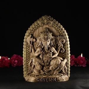 May include: A wooden carving of the Hindu deity Ganesha, with a golden frame and intricate details. The carving depicts Ganesha sitting on a throne with his elephant head and multiple arms. The inscription "GANESHA" is visible at the bottom of the carving.