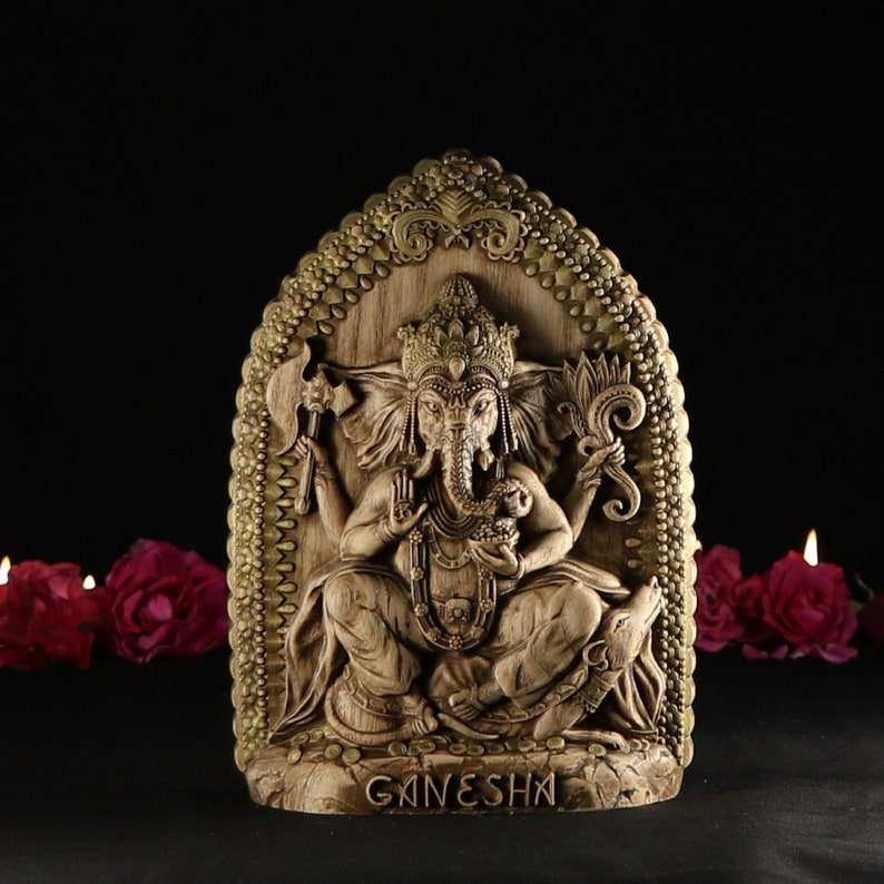 May include: A wooden carving of the Hindu deity Ganesha, with a golden frame and intricate details. The carving depicts Ganesha sitting on a throne with his elephant head and multiple arms. The inscription "GANESHA" is visible at the bottom of the carving.